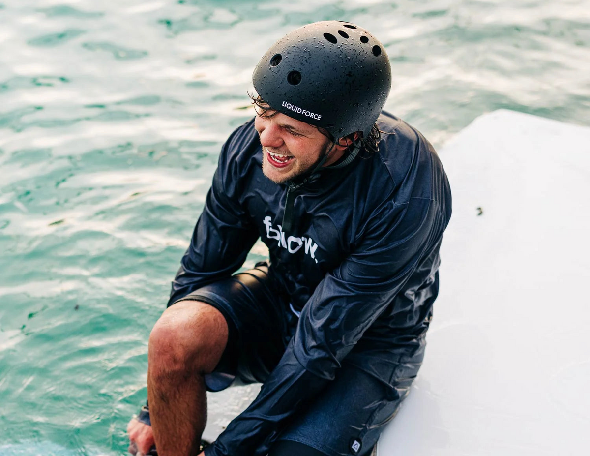 A man wearing a black Liquid Force helmet and a black wet-look rash guard sits on the edge of a white boat in turquoise water, smiling.