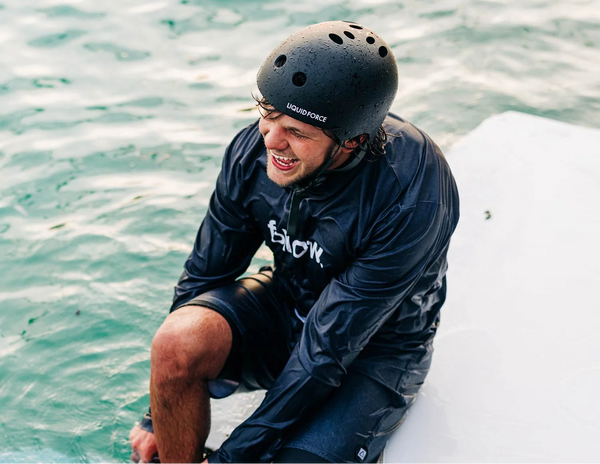 A man wearing a black Liquid Force helmet and a black wet-look rash guard sits on the edge of a white boat in turquoise water, smiling.